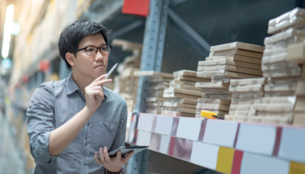 Young Asian man worker doing stocktaking of product in cardboard box on shelves in warehouse by using digital tablet and pen. Physical inventory count concept