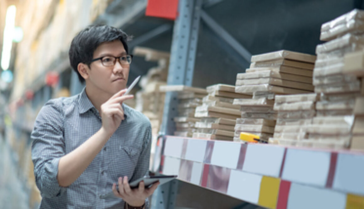 Young Asian man worker doing stocktaking of product in cardboard box on shelves in warehouse by using digital tablet and pen. Physical inventory count concept