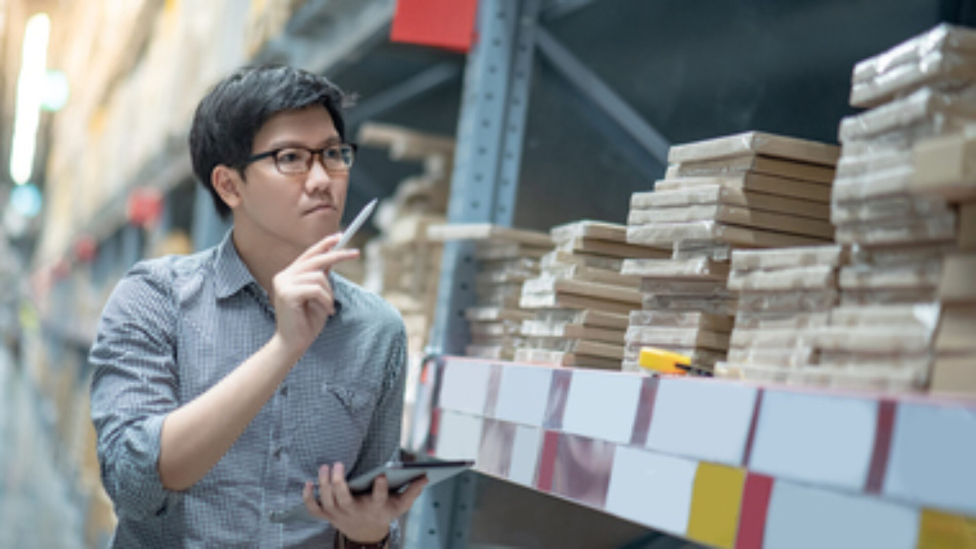 Young Asian man worker doing stocktaking of product in cardboard box on shelves in warehouse by using digital tablet and pen. Physical inventory count concept