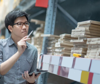 Young Asian man worker doing stocktaking of product in cardboard box on shelves in warehouse by using digital tablet and pen. Physical inventory count concept