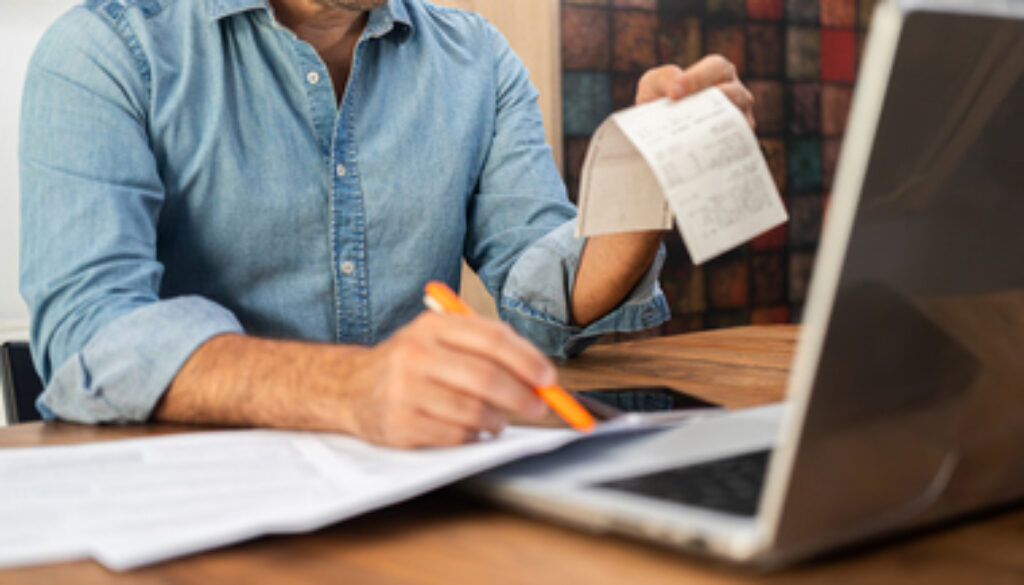 Middle-aged man calculating taxes at a wooden desk with a laptop and holding receipts. Personal finance management and accounting concept during tax season.