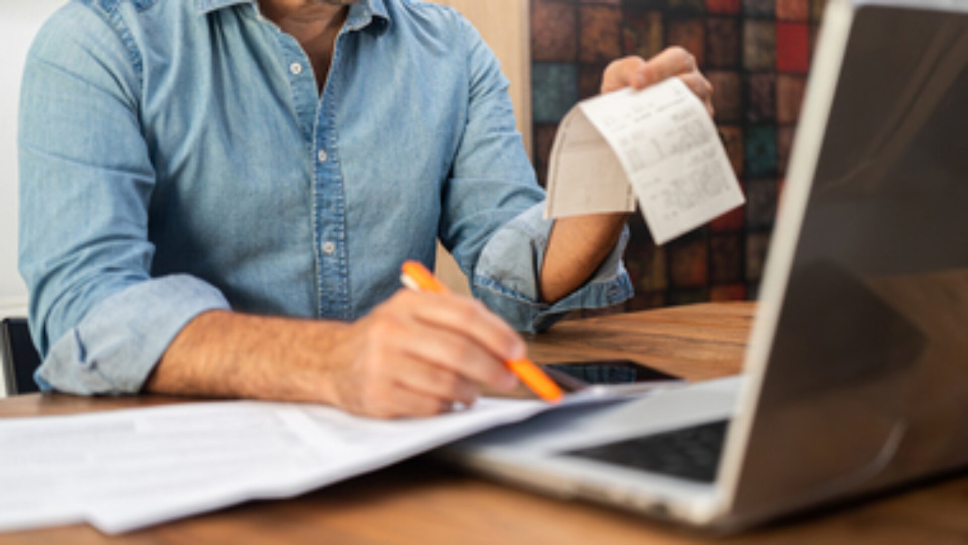 Middle-aged man calculating taxes at a wooden desk with a laptop and holding receipts. Personal finance management and accounting concept during tax season.