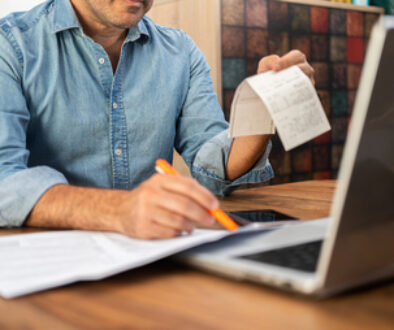 Middle-aged man calculating taxes at a wooden desk with a laptop and holding receipts. Personal finance management and accounting concept during tax season.