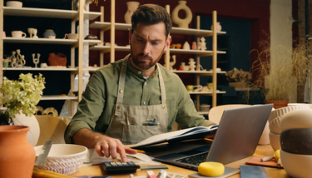 Serious frowned Caucasian man male small business owner businessman potter craftsman counting expense payment calculator taxes working laptop look notebook writing accounting indoors pottery workshop