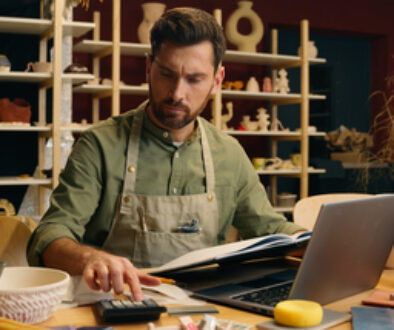 Serious frowned Caucasian man male small business owner businessman potter craftsman counting expense payment calculator taxes working laptop look notebook writing accounting indoors pottery workshop